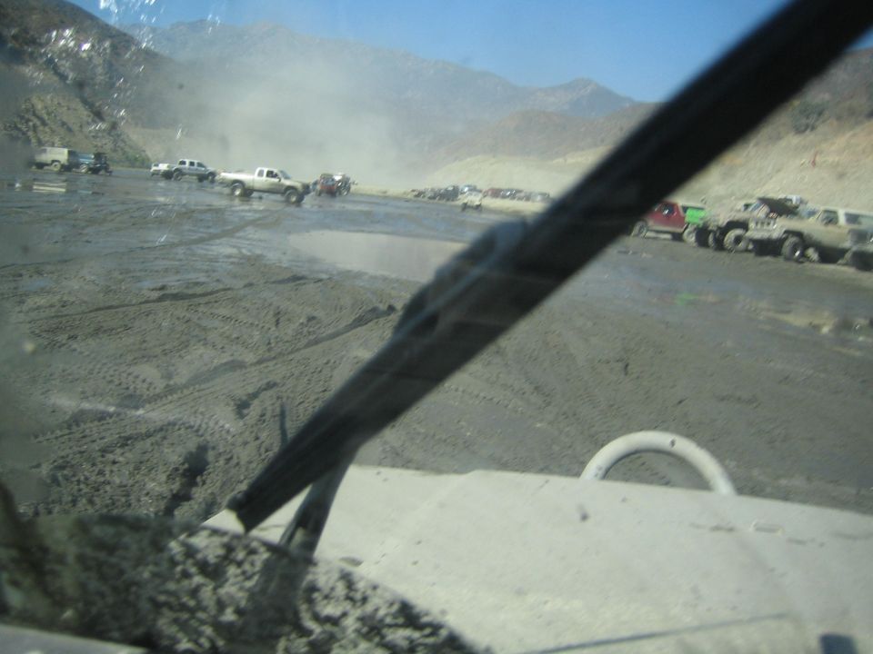 View from inside a vehicle looking out at several trucks and SUVs in a muddy, dusty off-road area with mountains in the background
