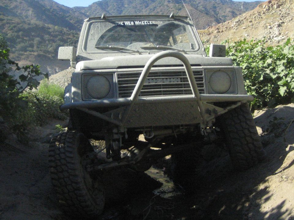 Suzuki Samurai on a dirt trail, front view. It has a custom bumper and is covered in mud