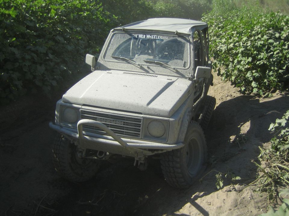 Suzuki Samurai driving on a dirt path with vegetation on both sides. Text on windshield reads 'THE WEB WHEELER'
