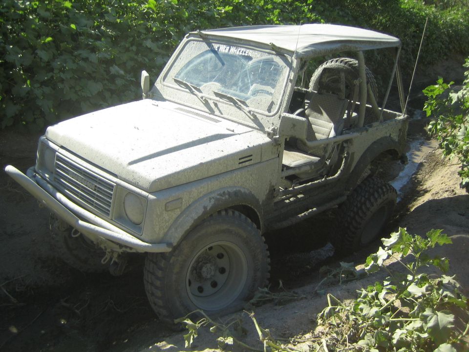 Mud-covered Suzuki Samurai off-roading in a muddy ditch, front-left three-quarter view