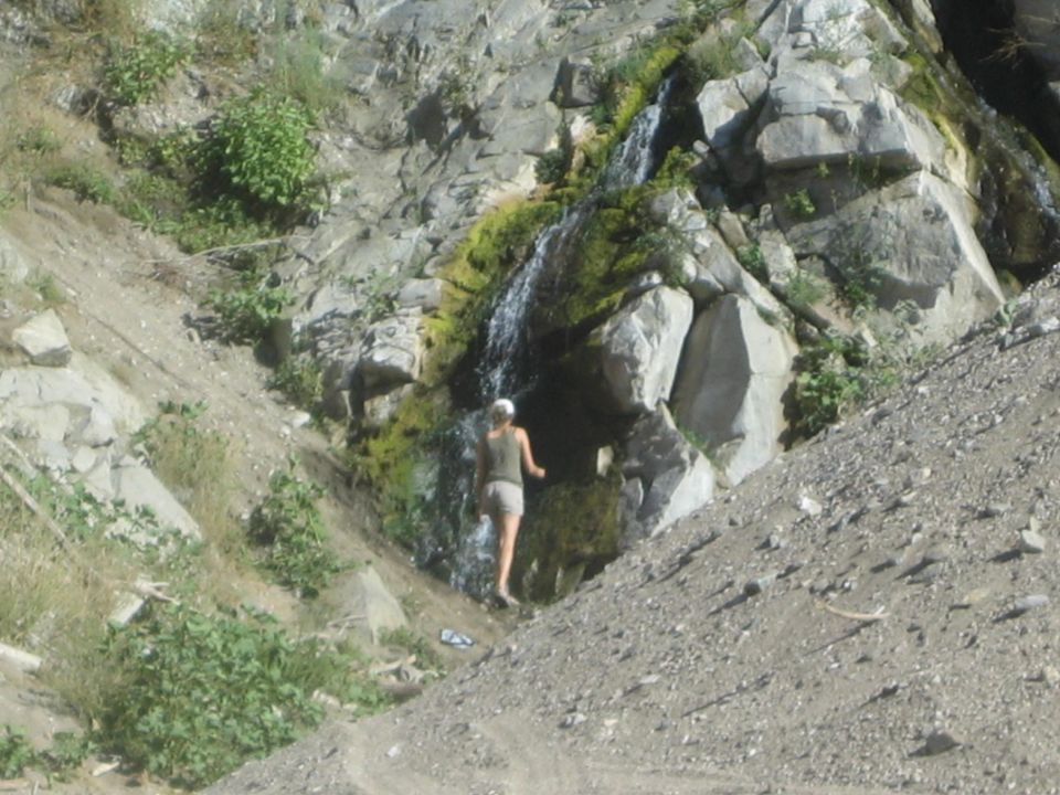 Woman in shorts and a tank top walks near a small waterfall cascading down a rocky, vegetated slope