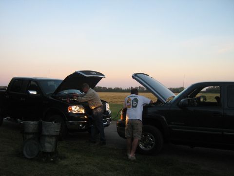 1995 Ford Bronco - Two trucks are parked in a field at dusk, with their hoods open as two people attend to them.