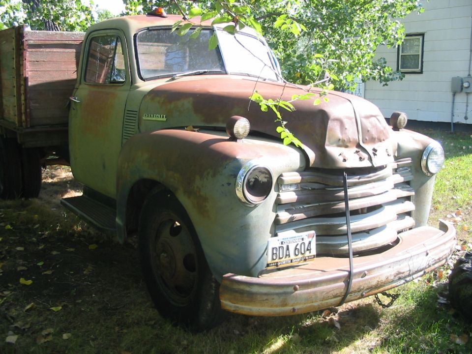 Rusty green 1950s Chevrolet Advance Design stake-bed truck parked in a yard, front three-quarter view