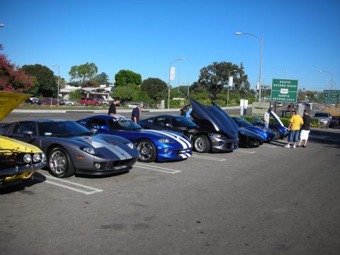 1998 Ford Crown Victoria - lineup of exotic cars including a silver-striped Ford GT and blue Dodge Vipers parked at a car show near a freeway sign.