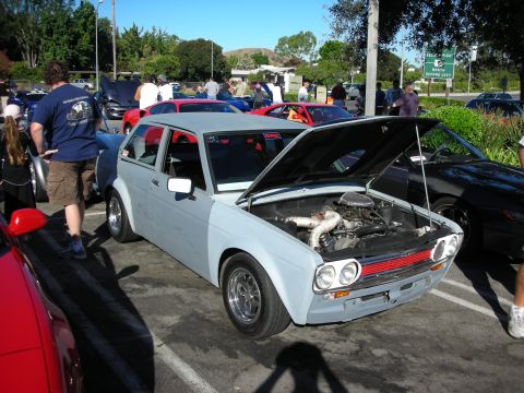 1998 Ford Crown Victoria - light blue vintage Datsun 510-style two-door coupe sits hood-up with a turbo intake visible at an outdoor car show.