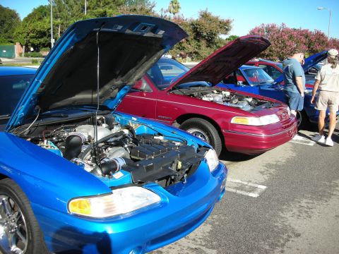 1998 Ford Crown Victoria - blue car and a magenta Ford sedan sit with hoods open at a sunny parking-lot car show with spectators nearby.