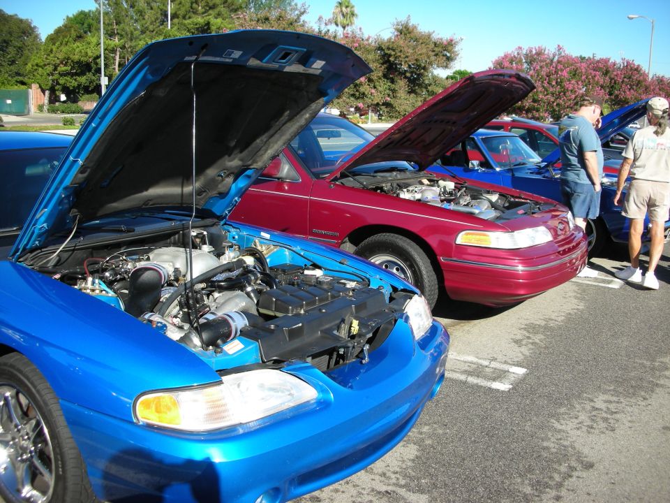 Blue car and magenta sedan with open hoods parked at an outdoor car show