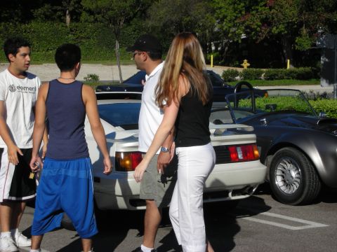 1998 Ford Crown Victoria - group of people stand chatting beside a silver sports car and a dark roadster in a sunny parking lot.