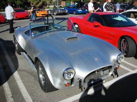 1998 Ford Crown Victoria - unpainted silver Shelby Cobra-style roadster with roll bar sits beside a red Corvette at a car show.
