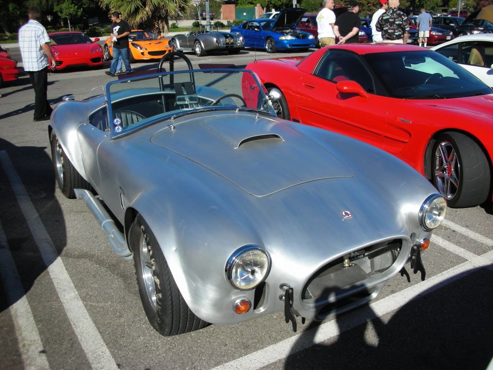 Bare-metal Shelby Cobra replica roadster parked beside a red Corvette
