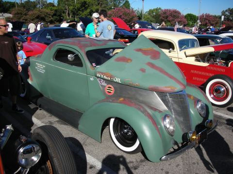 1998 Ford Crown Victoria - weathered green vintage coupe hot rod with whitewall tires and Wynn-Oil lettering parked at a car show.