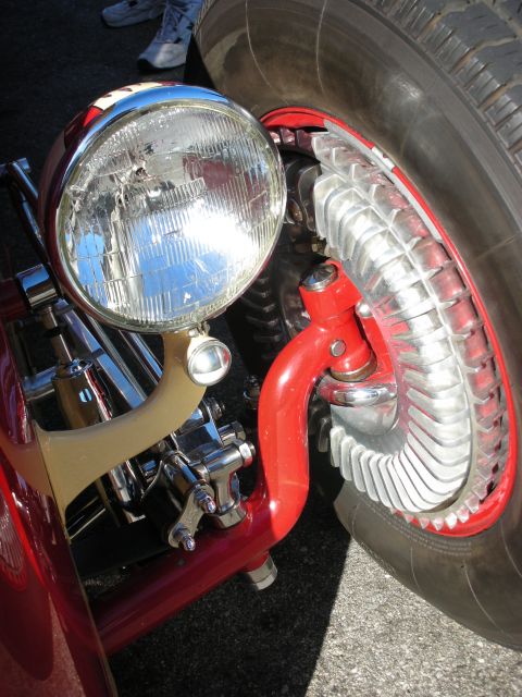 1998 Ford Crown Victoria - Close-up of a chrome headlight and large finned brake drum on the red front suspension of a hot rod.