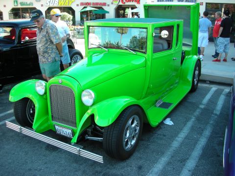 1998 Ford Crown Victoria - bright green vintage-style hot rod pickup with chrome grille and modern wheels parked on a street lined with shops and onlookers.