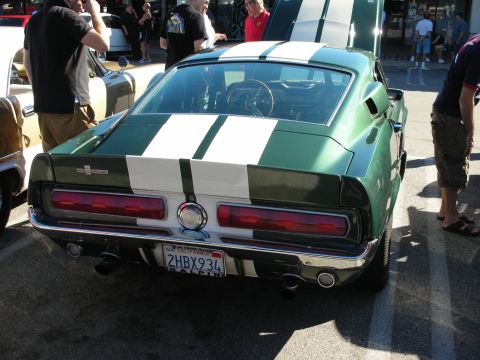1998 Ford Crown Victoria - Rear view of a green fastback muscle car with twin white racing stripes and dual exhaust, parked among onlookers at a show.