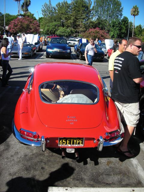 1998 Ford Crown Victoria - Rear of a red Jaguar E-Type coupe with chrome bumpers and a California '65 ETYPE' plate, parked among cars and people.