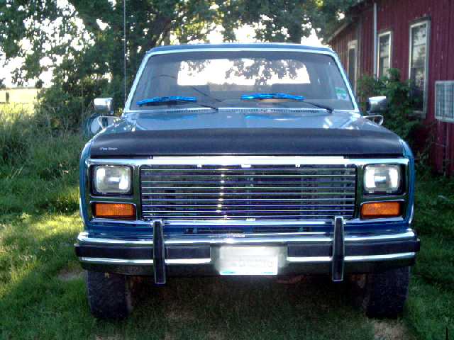 Front view of blue Ford Bronco with black hood deflector, chrome grille and push bar, parked on grass by red barn