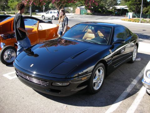 1998 Ford Crown Victoria - Black Ferrari 456 grand tourer parked at a car show, front three-quarter view with tan interior, beside an orange hot rod.