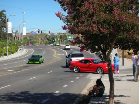 1998 Ford Crown Victoria - Roadside scene showing a red coupe parked on a flatbed-lined street while a green vintage muscle car drives along the curving road.