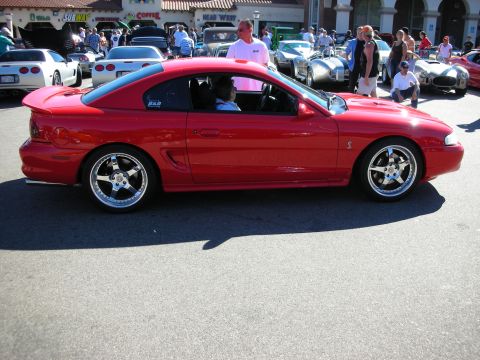 1998 Ford Crown Victoria - Side profile of a red Ford Mustang SVT Cobra coupe on chrome five-spoke wheels, parked among other cars at a crowded car show.