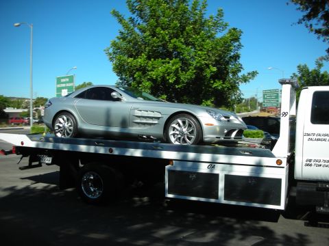 1998 Ford Crown Victoria - Silver Mercedes-Benz SLR McLaren coupe loaded sideways on a flatbed tow truck parked along a tree-lined street.