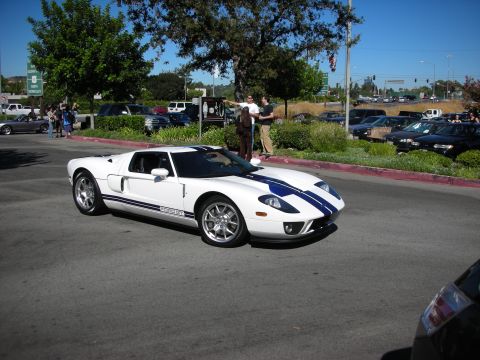 1998 Ford Crown Victoria - White Ford GT supercar with twin blue racing stripes driving through a tree-lined parking lot as spectators watch at a car show.