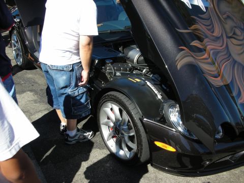1998 Ford Crown Victoria - Black sports car with its hood raised, flame-style airbrush artwork visible, exposing the engine and a chrome wheel as onlookers stand by.