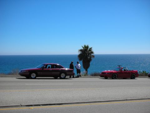 1998 Ford Crown Victoria - dark red sedan and a red convertible parked along a coastal highway with two people standing between them and the ocean behind.