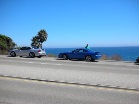 1998 Ford Crown Victoria - blue sport coupe and a silver car parked along a coastal highway with a person standing nearby and the ocean in the background.