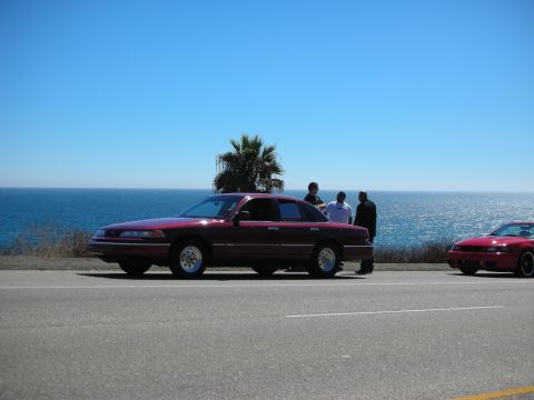 1998 Ford Crown Victoria - dark red Ford sedan parked along a coastal highway with three people gathered beside it and the ocean in the background.