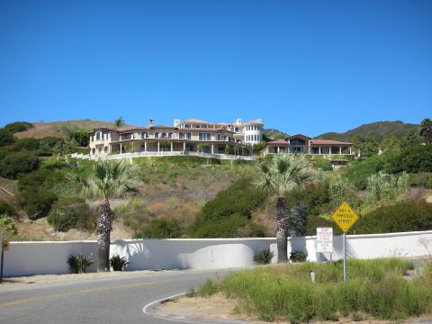 1998 Ford Crown Victoria - large multi-level mansion sits atop a green hillside above a white retaining wall, with palm trees and a yellow road sign in the foreground.