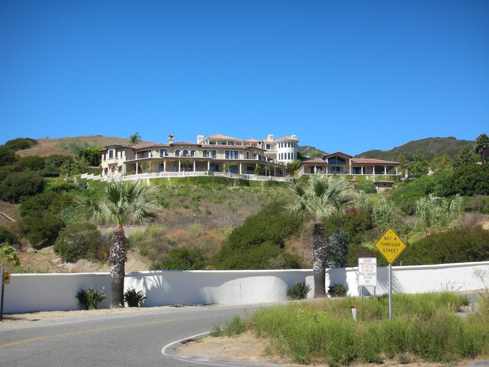 Large hillside mansion above a white wall with palm trees and a road sign