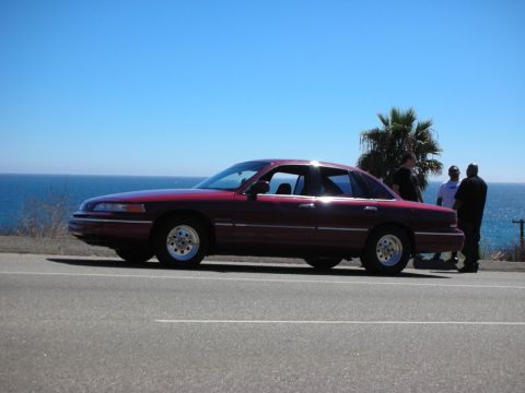 1992 Ford Crown Victoria - A maroon 1992 Ford Crown Victoria sedan is parked on a roadside overlooking the ocean with three people standing nearby.