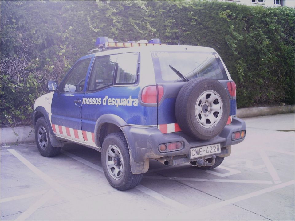 Rear view of a blue and white police SUV with 'mossos d'esquadra' text and red and white stripes. Spare tire mounted on the back