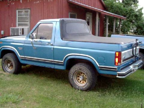 1984 Ford Bronco - A blue Ford Bronco with a soft top is parked on grass in front of a red building, with another vehicle visible behind it.
