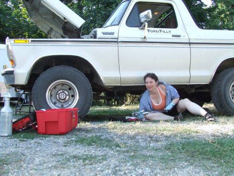 1979 Ford Bronco - A woman works on a white 1979 Ford Bronco, which has its hood open and is parked outdoors.