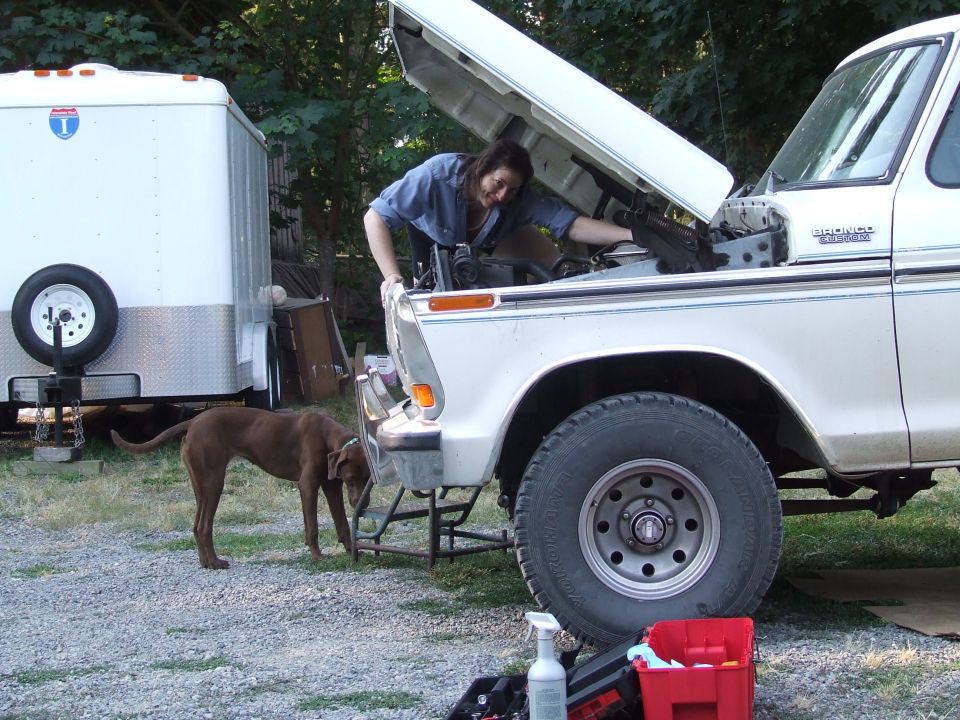 Person working under the open hood of a white and tan Ford Bronco Custom in a gravel driveway
