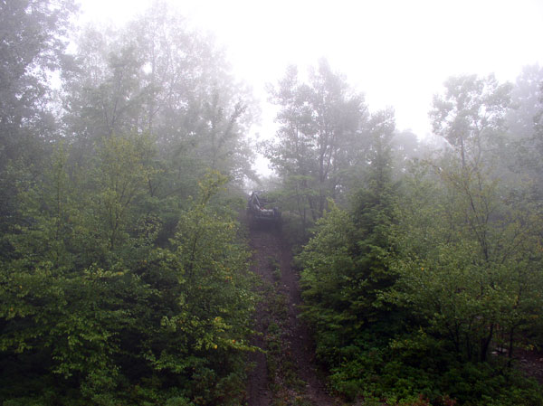 Dark 4x4 cresting a rutted dirt trail hill in heavy fog, seen from a distance