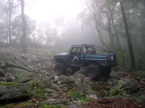 1988 Ford Bronco - Off-road SUV with roll cage driving through a rocky, wooded area on a foggy day.