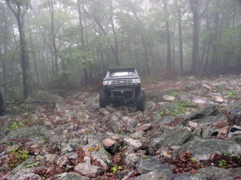 1988 Ford Bronco - Head-on front view of a lifted dark full-size Ford Bronco with the top removed, roll bar visible, crawling over a rocky boulder field on a…