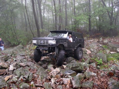 1988 Ford Bronco - A black 1988 Ford Bronco tackles a challenging rocky path in a wooded area, with a driver behind the wheel.