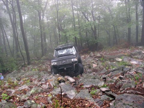 1988 Ford Bronco - Wide shot of a lifted black Ford 4x4 working down a rain-soaked boulder-strewn trail through foggy hardwood forest. The truck's bumperless…