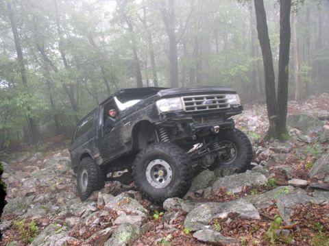 1988 Ford Bronco - A lifted black Ford Bronco crawls a wet boulder field in foggy woods, front axle articulated with one tire climbing a rock and the opposite…