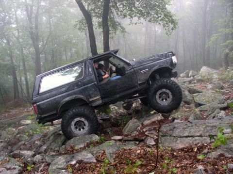 1988 Ford Bronco - Side view of a mud-splattered black Ford Bronco climbing a steep rock face in foggy woods, front end pitched up with the driver visible thr…