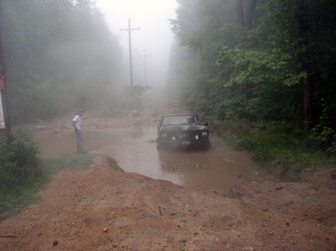 1988 Ford Bronco - A 1988 Ford Bronco is stuck in a muddy section of a dirt road surrounded by trees.