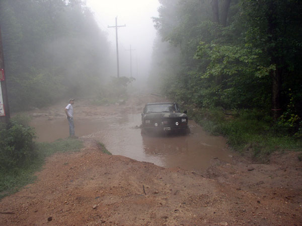 1988 Ford Bronco stuck in deep mud on a wooded dirt road with a person standing nearby