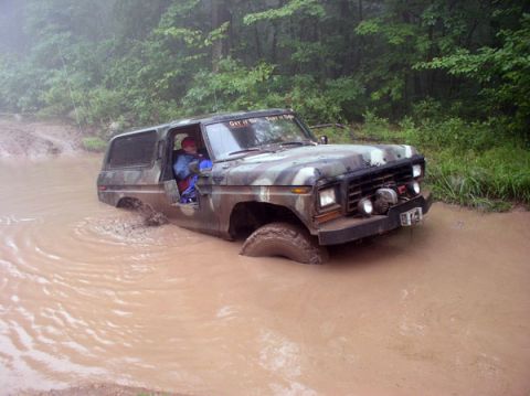 1988 Ford Bronco - A doorless camouflage-painted full-size Ford 4x4 wades through a muddy brown water hole on a wooded trail, water up over the tires and rock…