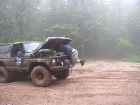 1988 Ford Bronco - A man works on the engine of a camouflaged Ford Bronco on a dirt track in a foggy forest.