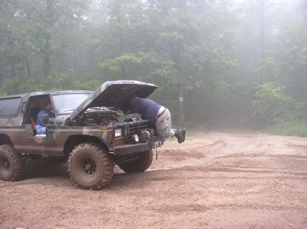 Camouflaged Ford Bronco with hood open, man working on engine, off-road tires, dirt track