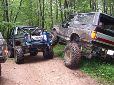 1988 Ford Bronco - Two heavily modified off-road vehicles navigate a trail in a wooded area.
