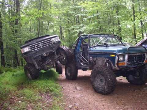 1988 Ford Bronco - Two heavily modified Ford trucks, one blue and one black, navigate a wooded trail.